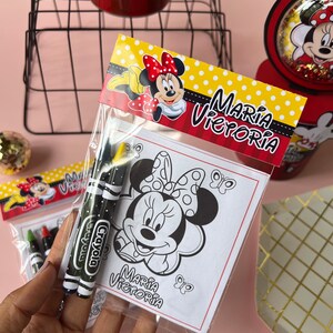 May include: A clear plastic bag filled with coloring supplies for a birthday party. The bag contains two black and green crayons with the word "Crayola" printed on them. The bag also contains a coloring page featuring a black and white illustration of Minnie Mouse with the text "Maria Victoria" printed below.