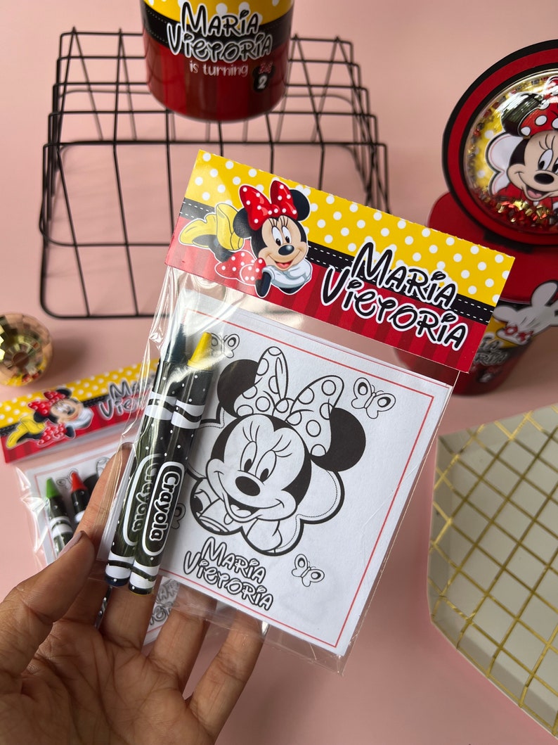 May include: A clear plastic bag filled with coloring supplies for a birthday party. The bag contains two black and green crayons with the word "Crayola" printed on them. The bag also contains a coloring page featuring a black and white illustration of Minnie Mouse with the text "Maria Victoria" printed below.