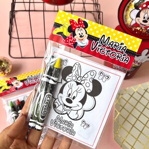 May include: A clear plastic bag filled with coloring supplies for a birthday party. The bag contains two crayons and a coloring page featuring a black and white image of Minnie Mouse. The coloring page has the text "Maria Victoria" at the bottom.