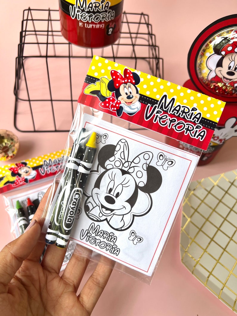 May include: A clear plastic bag filled with coloring supplies for a birthday party. The bag contains two crayons and a coloring page featuring a black and white image of Minnie Mouse. The coloring page has the text "Maria Victoria" at the bottom.