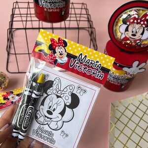 May include: A clear plastic bag filled with coloring supplies for a birthday party. The bag contains two black and red crayons and a coloring page featuring a black and white illustration of Minnie Mouse. The coloring page has the text "Maria Victoria" at the bottom.