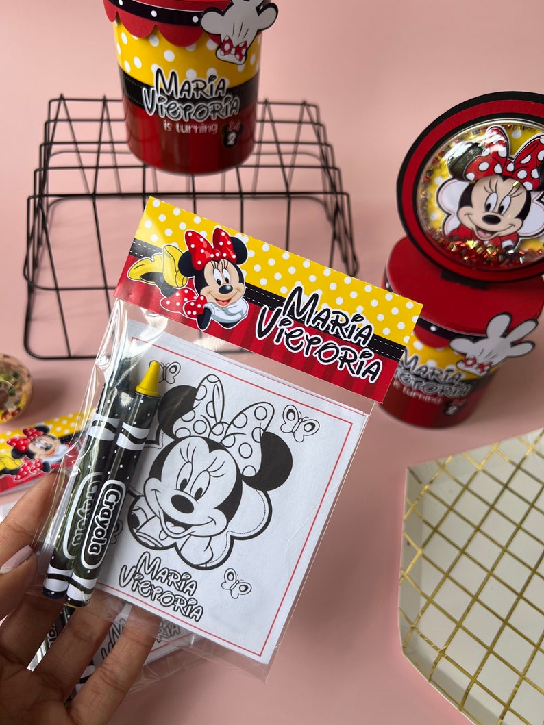 May include: A clear plastic bag filled with coloring supplies for a birthday party. The bag contains two black and red crayons and a coloring page featuring a black and white illustration of Minnie Mouse. The coloring page has the text "Maria Victoria" at the bottom.