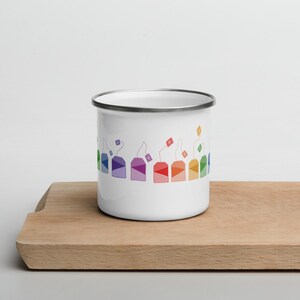 Può includere: Una tazza di smalto bianco con un motivo arcobaleno di bustine di tè attorno al bordo. Le bustine di tè sono di diversi colori e hanno dei lacci che pendono.