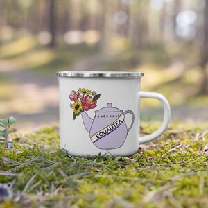 Può includere: Una tazza di smalto bianco con una teiera viola con un nastro giallo e viola che dice "Equalitea" e un mazzo di fiori nel beccuccio. La tazza è appoggiata su un letto di muschio verde.