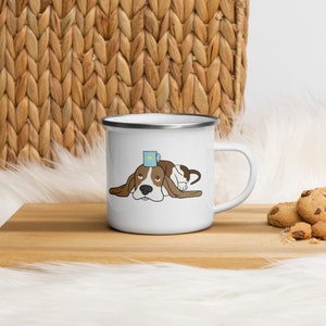 Pode incluir: Caneca de esmalte branco com uma ilustração de desenho animado de um basset hound com uma caneca azul na cabeça. O cão está deitado com a cabeça apoiada nas patas.