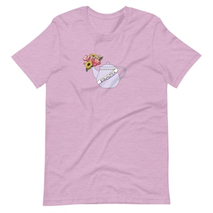 Op de afbeelding: Een lichtroze T-shirt met een cartoonillustratie van een theepot met bloemen en een band met de tekst "Equalitea".