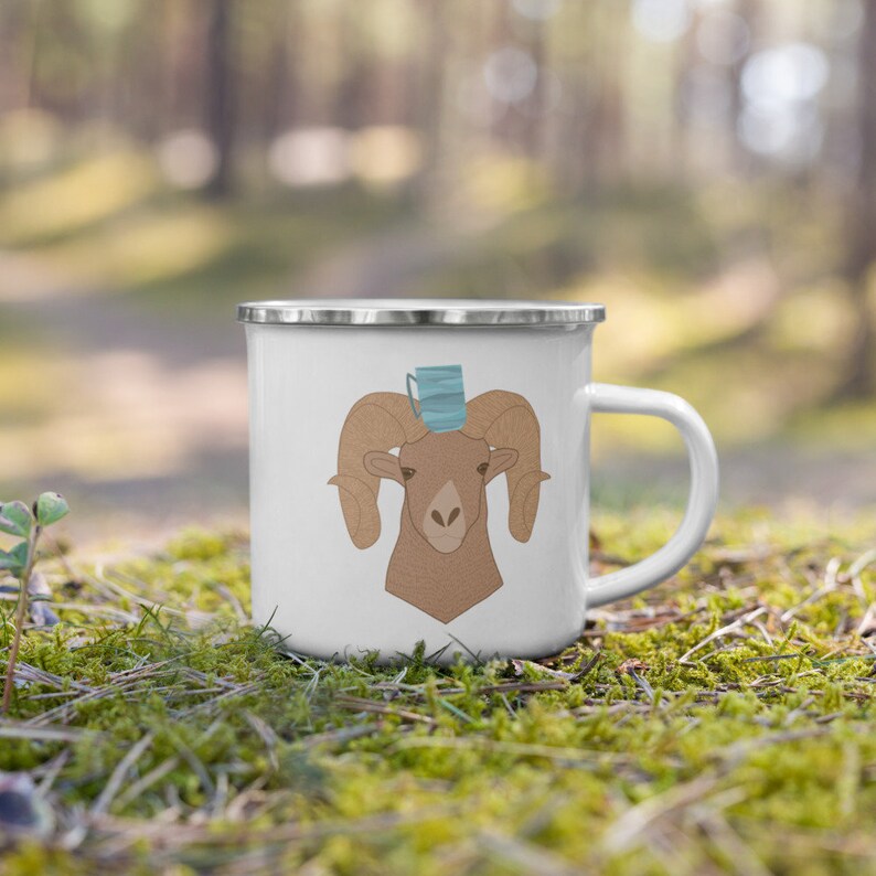 May include: White enamel camping mug with a brown ram head illustration wearing a blue face mask. The mug has a silver rim and handle.