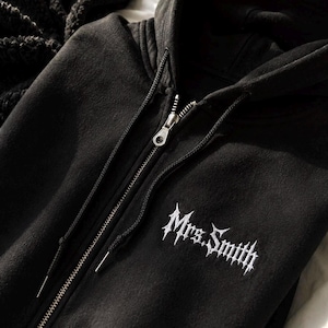 May include: A black zip-up hoodie featuring a hood with black drawstrings and a silver metal zipper pull. The chest area displays the text Mrs. Smith embroidered in a white, stylized, gothic-inspired font with sharp, pointed edges.