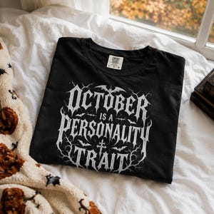 May include: A black short-sleeve t-shirt featuring white graphic text that reads OCTOBER IS A PERSONALITY TRAIT. The design includes intricate branches, small bats, and a cross symbol. The shirt has a crew neckline and a Comfort Colors brand label.