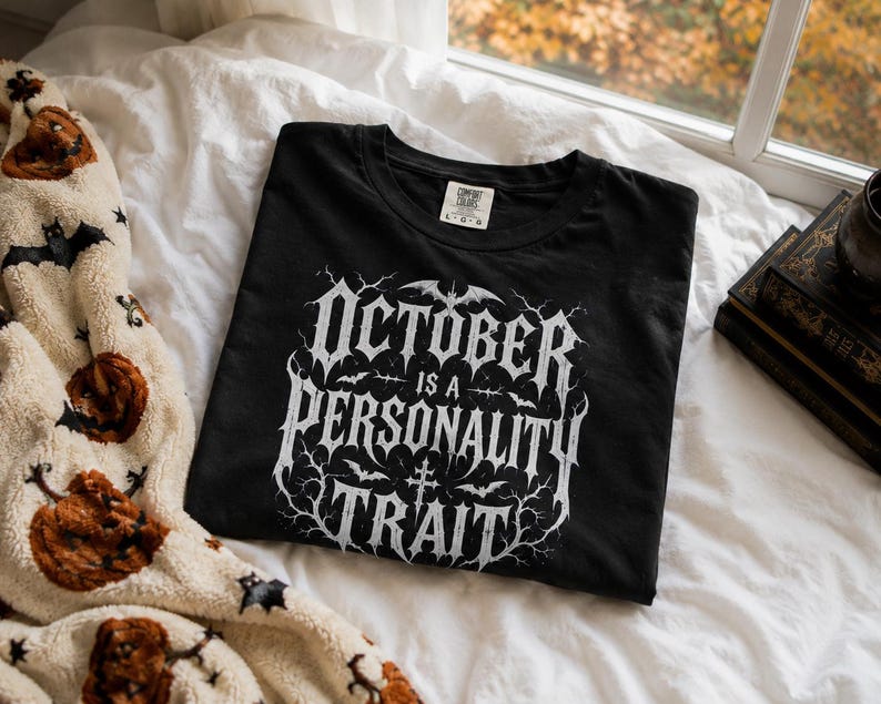 May include: A black short-sleeve t-shirt featuring white graphic text that reads OCTOBER IS A PERSONALITY TRAIT. The design includes intricate branches, small bats, and a cross symbol. The shirt has a crew neckline and a Comfort Colors brand label.