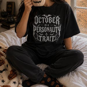 May include: A black short-sleeved t-shirt featuring white gothic-style text that reads OCTOBER IS A PERSONALITY TRAIT, surrounded by illustrations of bats and bare tree branches. The shirt is paired with black sweatpants and black pumpkin-patterned socks.