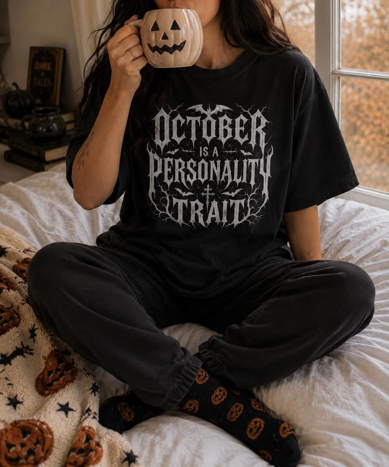 May include: A black short-sleeved t-shirt featuring white gothic-style text that reads OCTOBER IS A PERSONALITY TRAIT, surrounded by illustrations of bats and bare tree branches. The shirt is paired with black sweatpants and black pumpkin-patterned socks.