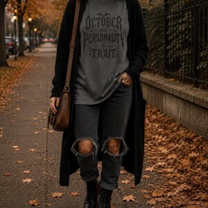 May include: A charcoal gray t-shirt with the black text "OCTOBER IS A PERSONALITY TRAIT" in a gothic font, worn under a long black open-front cardigan, paired with black distressed denim jeans featuring knee rips and black lace-up leather boots.