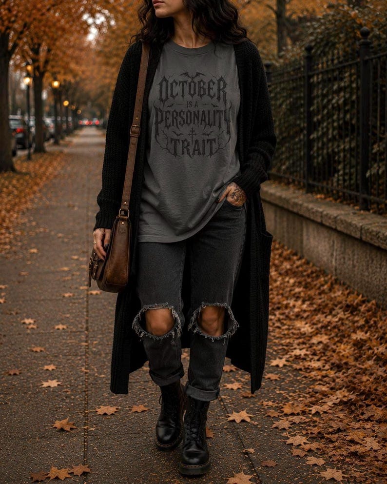 May include: A charcoal gray t-shirt with the black text "OCTOBER IS A PERSONALITY TRAIT" in a gothic font, worn under a long black open-front cardigan, paired with black distressed denim jeans featuring knee rips and black lace-up leather boots.