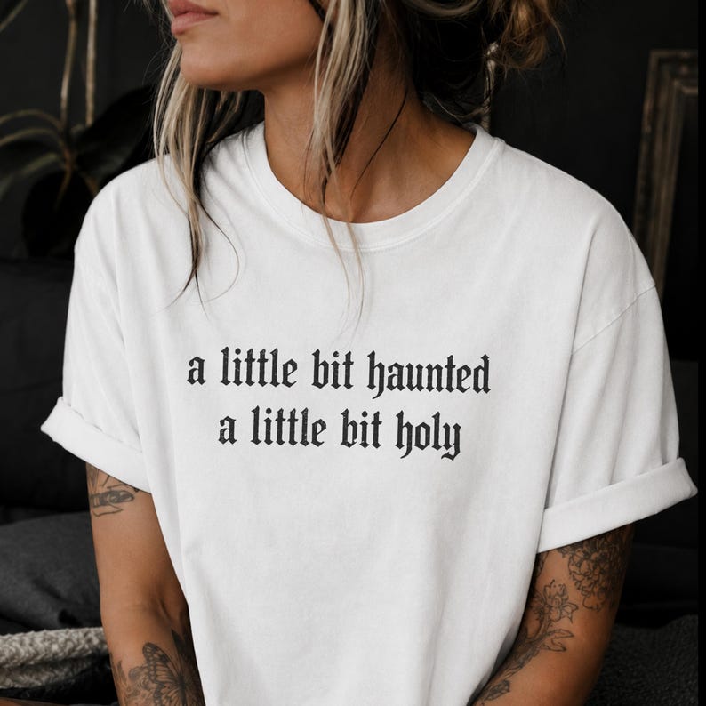 Op de afbeelding: Een wit T-shirt met korte mouwen, een ronde hals en opgerolde mouwen. Op het midden van de borst staat zwarte tekst in een gotisch lettertype die in twee regels luidt: a little bit haunted, a little bit holy.
