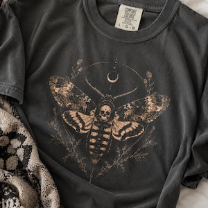 May include: A charcoal gray short-sleeve t-shirt featuring a graphic print of a moth with a skull pattern on its thorax, surrounded by a thin circular frame, botanical sprigs, and a crescent moon. The inner neck label reads COMFORT COLORS L-G-G.