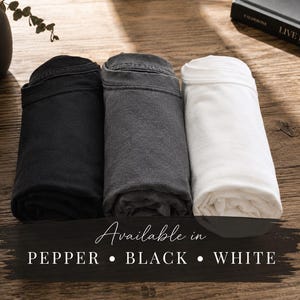 May include: Three rolled t-shirts in black, charcoal gray, and white. The text overlay reads: Available in PEPPER &bull; BLACK &bull; WHITE. The shirts are arranged side-by-side, showcasing the distinct color variations of the fabric in a simple, clean presentation.