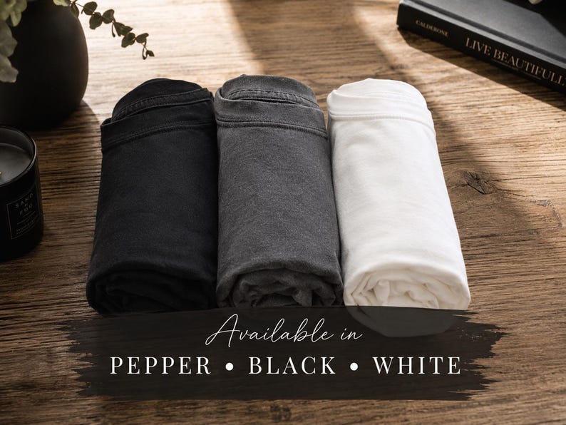 May include: Three rolled t-shirts in black, charcoal gray, and white. The text overlay reads: Available in PEPPER &bull; BLACK &bull; WHITE. The shirts are arranged side-by-side, showcasing the distinct color variations of the fabric in a simple, clean presentation.