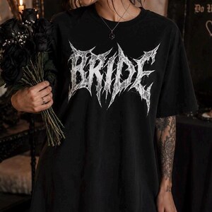 May include: A black short-sleeved t-shirt featuring the word BRIDE printed in a white, distressed, jagged, metal-style font across the chest. The shirt has a crew neckline and a loose, relaxed fit, made of a solid black fabric.