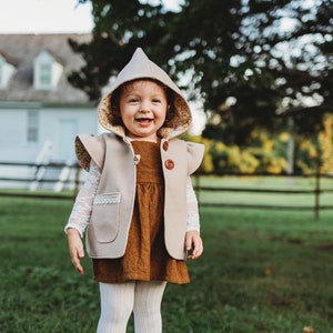 May include: A young child wearing a brown corduroy dress with a beige hooded vest and white tights. The vest has a floral lining and lace trim. The child is smiling and standing on a stone surface.