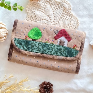 Peut inclure: Un portefeuille matelassé beige avec un motif de paysage fantaisiste. Le rabat présente un arbre vert, une maison avec un toit rouge et une bordure florale verte. Le portefeuille a une bordure à carreaux marron et une fermeture à bouton.