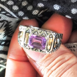 May include: A silver ring with a rectangular, purple amethyst gemstone. The ring has gold accents and a detailed, patterned design around the band. The ring is held in a hand.