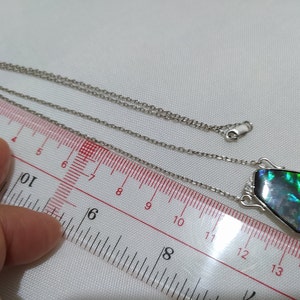 May include: A silver chain necklace with a pendant featuring a dark green, blue, and iridescent gemstone. The pendant is about 2.5 inches long and is measured against a ruler.
