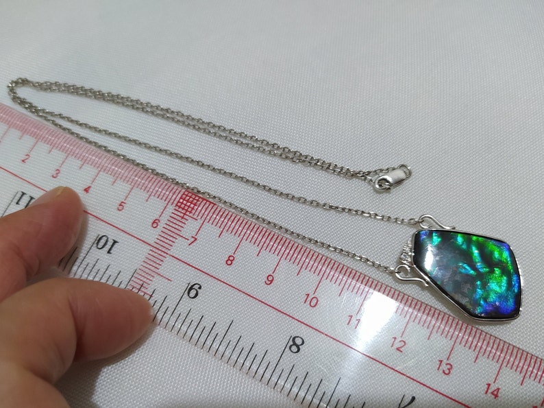 May include: A silver chain necklace with a pendant featuring a dark green, blue, and iridescent gemstone. The pendant is about 2.5 inches long and is measured against a ruler.