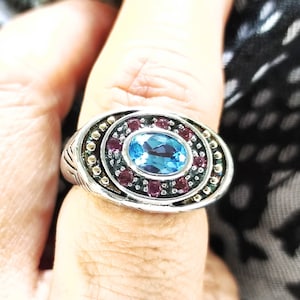 May include: Silver ring featuring an oval, light blue gemstone at the center, surrounded by smaller, round, dark red stones. The ring has a detailed, textured band with a decorative pattern. The ring is a piece of jewelry.