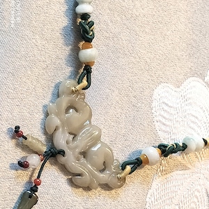 May include: A jade pendant necklace with a green jade carving of a floral design. The necklace is made with green, brown, and white beads and cord.
