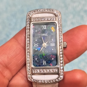 May include: Rectangular wristwatch with a white leather band and a silver-tone case. The watch face features an opal stone with Roman numerals and the word "OPAL". The case is encrusted with small, clear gemstones.
