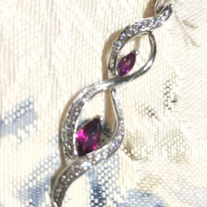 May include: A silver pendant with a swirling design and two purple gemstones. The pendant is set with small clear stones.