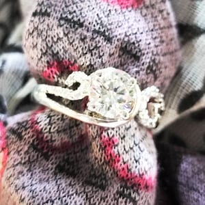 May include: A silver ring with a large, clear, round gemstone surrounded by smaller gemstones. The ring is on a patterned fabric.