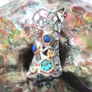 May include: A silver and gold pendant with a geometric design and colorful gemstones. The pendant features blue, yellow, and turquoise gemstones set in a honeycomb pattern.