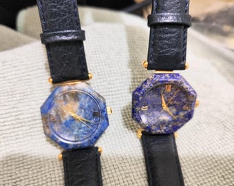 May include: Two wristwatches with octagonal faces made of blue lapis lazuli stone, featuring gold-tone hands and Roman numeral markers. Each watch has a textured black leather strap with gold-tone hardware connecting the strap to the watch case.