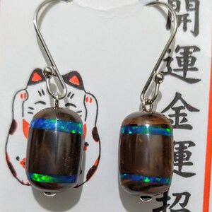 May include: A pair of silver dangle earrings with brown and black oval-shaped stones. The stones have a blue and green iridescent sheen. The earrings are hanging from a white card with Japanese characters that read "開運金運招".