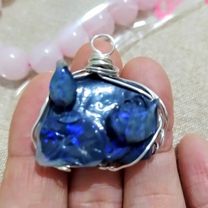 May include: A silver wire-wrapped pendant featuring a dark blue, iridescent opal carved into the shape of a pig's head.