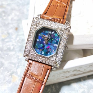 Puede incluir: Un reloj de forma cuadrada con una correa de cuero marrón. La esfera del reloj está rodeada por una fila de piedras transparentes y presenta una esfera de ópalo azul iridiscente con marcadores de hora en números romanos.