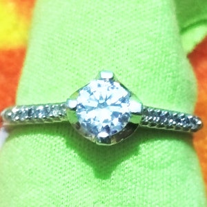 May include: A silver ring with a clear, round gemstone in the center. The ring has a textured band.