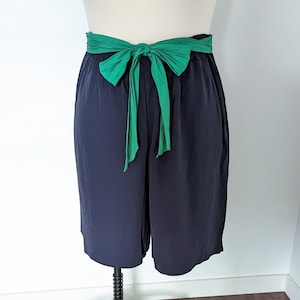 Könnte beinhalten: Marineblaue Shorts mit hoher Taille und breitem, grünem Schleifengürtel. Die Shorts werden auf einer Schaufensterpuppe vor weißem Hintergrund präsentiert. Die Schleife ist vorne gebunden und setzt einen Farbtupfer.