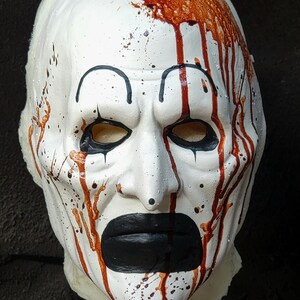 May include: A white mask with black eye outlines, a black mouth, and red blood-like streaks and splatters. The mask has a clown-like appearance, with a stern expression. The mask is likely for Halloween or costume use.