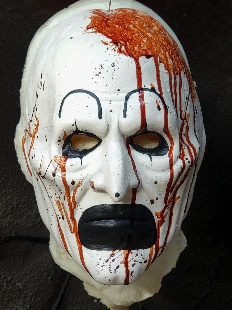 May include: A white mask with black eye outlines, a black mouth, and red blood-like streaks and splatters. The mask has a clown-like appearance, with a stern expression. The mask is likely for Halloween or costume use.