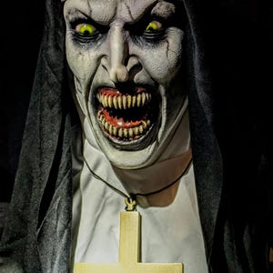May include: A close-up of a terrifying nun with glowing yellow eyes and a menacing grin. The nun is wearing a black habit and a white wimple. A gold cross hangs around the neck. The image is dark and foreboding, perfect for Halloween.