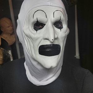 May include: A white clown mask with black accents, including eyebrows, teardrops, and a large mouth. The mask has a small black top hat. The person is wearing a black t-shirt with red text that reads "NIGHT OF THE".
