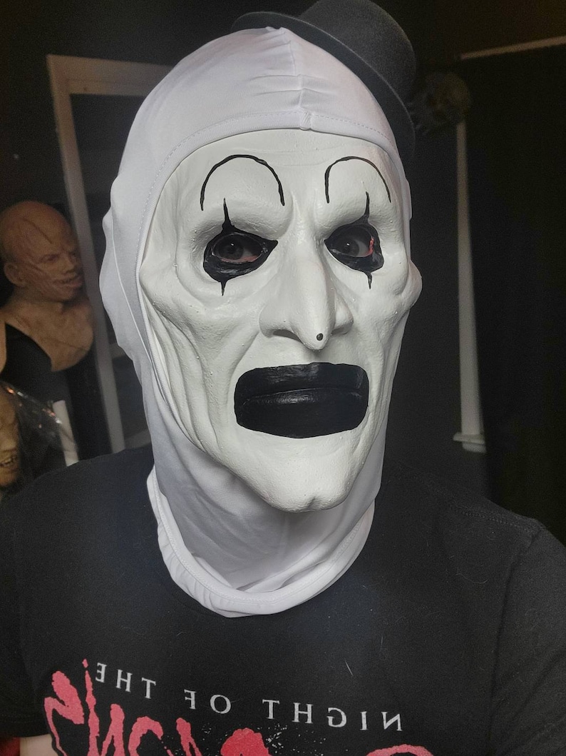 May include: A white clown mask with black accents, including eyebrows, teardrops, and a large mouth. The mask has a small black top hat. The person is wearing a black t-shirt with red text that reads "NIGHT OF THE".