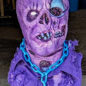 Puede incluir: Una máscara de zombi texturizada de color morado con aspecto desgastado, que presenta un ojo blanco y una cuenca ocular oscura y hueca. La máscara se combina con un collar de cadena de plástico azul brillante y una camisa morada deshilachada.
