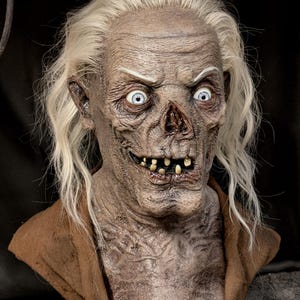 Puede incluir: Máscara detallada de una criatura muerta viviente en descomposición. La máscara presenta piel pálida, ojos hundidos, dientes expuestos y cabello largo y blanco. La máscara es marrón y beige con un cuello de tela marrón, ideal para Halloween.