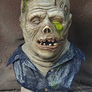 May include: A detailed bust of a zombie figure featuring textured, pale, decaying skin with green accents, a deep gash on the forehead, and one milky white eye. The figure wears a dark blue collared shirt with visible buttons and green slime.