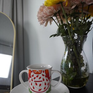 May include: A white ceramic two-handled mug and matching saucer set featuring a colorful graphic print of a stylized character wearing an apron with red strawberry motifs. A clear glass vase filled with yellow and pink chrysanthemum flowers.