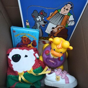 Op de afbeelding: Een verzameling kinderartikelen, waaronder een blauwe puzzel met figuren uit Lady en de Vagebond, een klein boekje getiteld "Bear Loves Weather", een rood-groene tas, een geel-paars Playskool-speelgoed en een witte schoen met roze accenten.